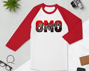 Puede incluir: Una camiseta de béisbol blanca y roja con un gráfico negro que dice "Elegua Omo" en una fuente estilizada. El gráfico tiene un degradado rojo, naranja y negro.