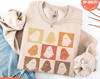 May include: Beige crew neck sweatshirt with a grid of nine ghost graphics in yellow, orange, and brown squares. The sweatshirt has a round neckline and long sleeves. The words "MORE COLOR OPTIONS" are in the bottom left corner. A silver necklace is visible.