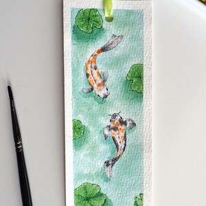 May include: A rectangular paper bookmark featuring a watercolor painting of two koi fish swimming in light green water with green lily pads. The bookmark has a light green ribbon attached at the top and a small black signature in the bottom corner.