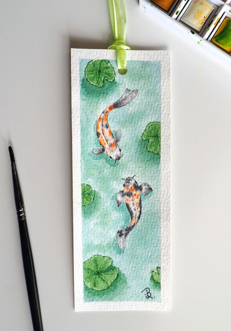 May include: A rectangular paper bookmark featuring a watercolor painting of two koi fish swimming in light green water with green lily pads. The bookmark has a light green ribbon attached at the top and a small black signature in the bottom corner.