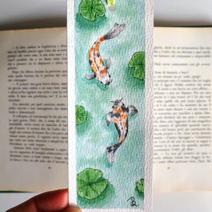 May include: A rectangular paper bookmark featuring a watercolor painting of two koi fish swimming in light green water with dark green lily pads. The bookmark has a light green ribbon attached at the top and a small black signature in the corner.