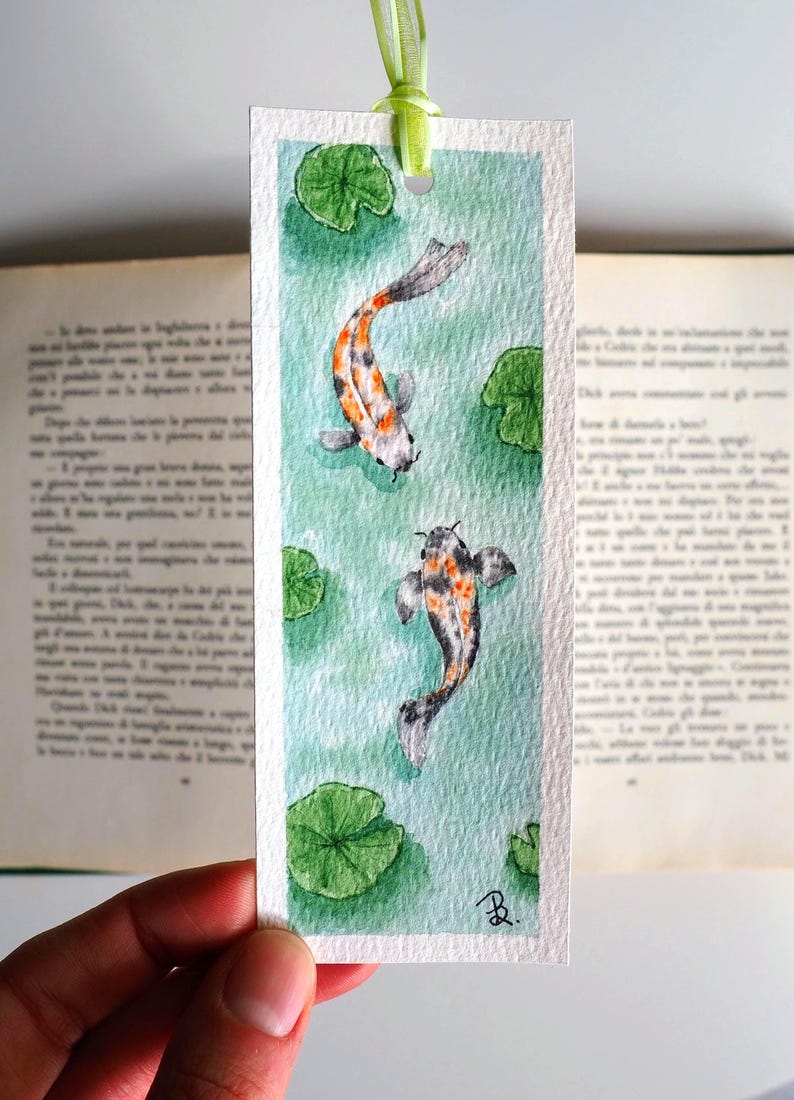May include: A rectangular paper bookmark featuring a watercolor painting of two koi fish swimming in light green water with dark green lily pads. The bookmark has a light green ribbon attached at the top and a small black signature in the corner.