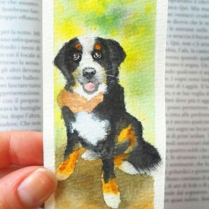 Può includere: Un segnalibro rettangolare in carta con un dipinto ad acquerello di un bovaro del bernese. Il cane ha il pelo nero, bianco e marrone, e il nome Olive è scritto in inchiostro nero in alto accanto a una piccola icona di un'impronta.