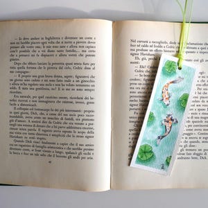 May include: A rectangular paper bookmark featuring a watercolor illustration of two koi fish swimming in green water with lily pads. The bookmark has a light green ribbon attached at the top and is placed inside an open book with printed text.