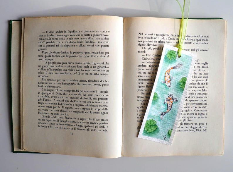 May include: A rectangular paper bookmark featuring a watercolor illustration of two koi fish swimming in green water with lily pads. The bookmark has a light green ribbon attached at the top and is placed inside an open book with printed text.