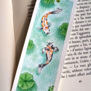 May include: A rectangular paper bookmark featuring a watercolor painting of two koi fish swimming in light green water with several green lily pads. The bookmark has a light green ribbon tied through a hole at the top and a small black signature.