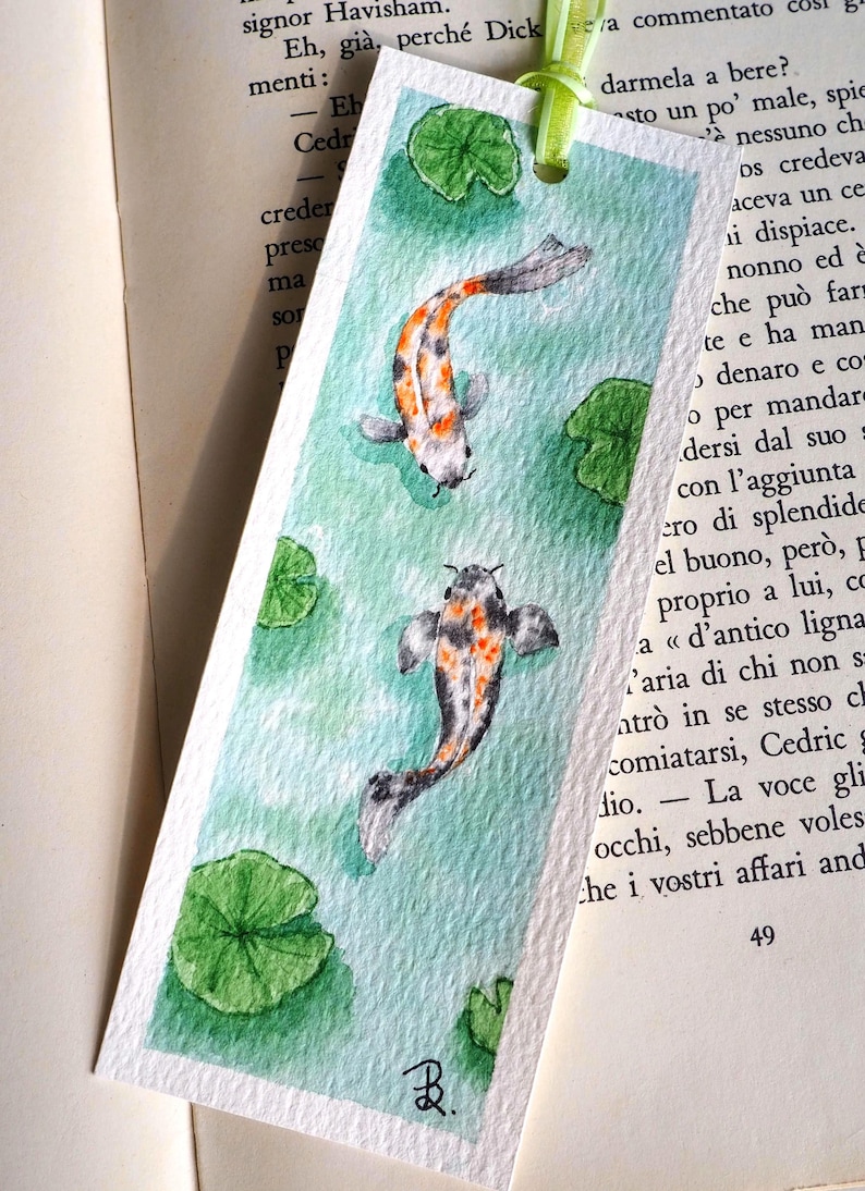 May include: A rectangular paper bookmark featuring a watercolor painting of two koi fish swimming in light green water with several green lily pads. The bookmark has a light green ribbon tied through a hole at the top and a small black signature.