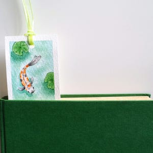 May include: A rectangular paper bookmark featuring a watercolor painting of a koi fish swimming among green lily pads in turquoise water. The bookmark has a hole at the top with a light green ribbon attached and is tucked into a dark green book.