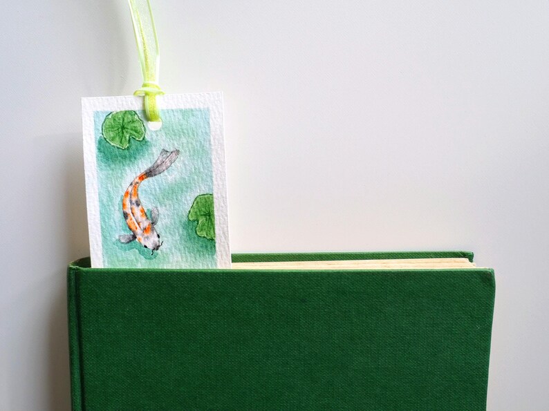 May include: A rectangular paper bookmark featuring a watercolor painting of a koi fish swimming among green lily pads in turquoise water. The bookmark has a hole at the top with a light green ribbon attached and is tucked into a dark green book.