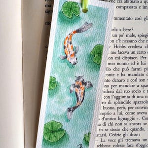 May include: A rectangular paper bookmark featuring a watercolor painting of two koi fish swimming in light green water with several green lily pads. The bookmark has a light green ribbon attached at the top and a small black signature in the corner.