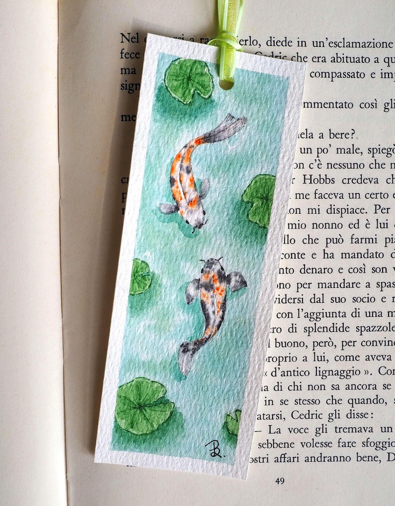 May include: A rectangular paper bookmark featuring a watercolor painting of two koi fish swimming in light green water with several green lily pads. The bookmark has a light green ribbon attached at the top and a small black signature in the corner.