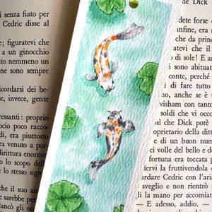 May include: A rectangular paper bookmark featuring a watercolor painting of two koi fish swimming in light green water with green lily pads. The bookmark has a light green ribbon tied through a hole at the top and a small black signature at the bottom.