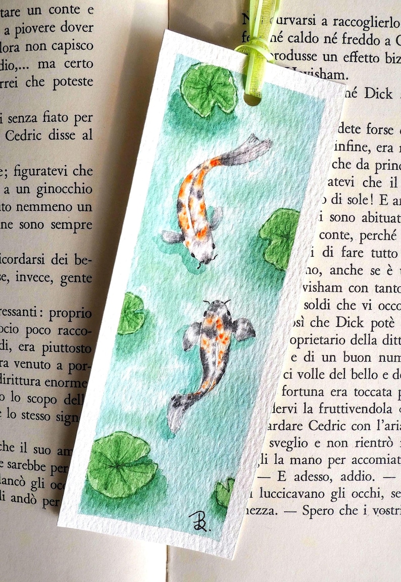May include: A rectangular paper bookmark featuring a watercolor painting of two koi fish swimming in light green water with green lily pads. The bookmark has a light green ribbon tied through a hole at the top and a small black signature at the bottom.