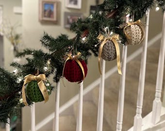 May include: Four crocheted Christmas ornaments hanging on a white railing with a garland of evergreen branches. The ornaments are decorated with gold ribbon bows and are red, green, beige, and brown.