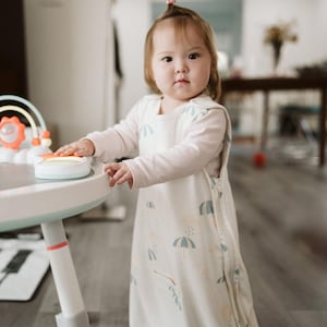 May include: A baby wearing a white sleep sack with a blue and yellow umbrella pattern. The baby is standing next to a white and light blue activity table with a toy on top. The baby is wearing a long-sleeved white shirt.