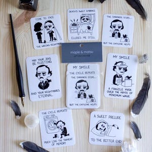 May include: A selection of square cards with black and white illustrations and text, featuring cartoon figures and phrases like "Time to Face" and "My Smile." The cards are arranged on a light wooden surface, with feathers, ink, and crystals.