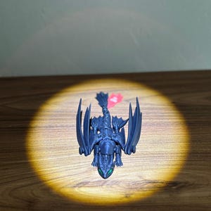 May include: A dark blue dragon figurine with outstretched wings and green eyes. The dragon is centered in a spotlight on a wooden surface. A red and pink object is visible near the dragon.