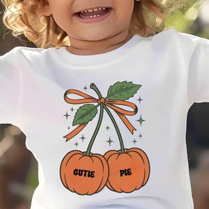 May include: White t-shirt featuring a whimsical design of two orange pumpkins with the words "CUTIE PIE" printed on them. The pumpkins are connected by a green stem, leaves, and an orange bow. The design is surrounded by sparkling stars.