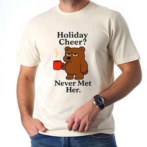 May include: Cream-colored t-shirt featuring a grumpy cartoon bear holding a red mug. The shirt has the text "Holiday Cheer? Never Met Her." The bear has a disgruntled expression. The shirt is short-sleeved.