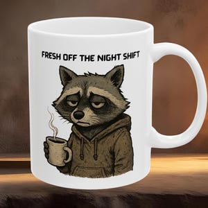 Op de afbeelding: Witte keramische koffiemok met een illustratie van een vermoeide wasbeer die een koffiekopje vasthoudt. De wasbeer draagt een bruine hoodie. De mok heeft de tekst "FRESH OFF THE NIGHT SHIFT" in zwarte letters.