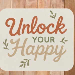 May include: A rectangular sign with rounded corners features the words "Unlock Your Happy" in orange and beige script. Small green leaf accents are placed around the text. The sign is set against a wooden background.