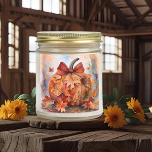 Könnte beinhalten: Ein Glas-Kerzenglas mit goldenem Deckel, mit einem Aquarelldesign eines Kürbisses, der mit Herbstblättern und einer roten Schleife verziert ist. Die Kerze steht auf einer rustikalen Holzoberfläche, mit orangefarbenen Blumen und Grün.