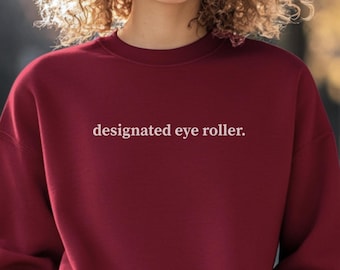 May include: A burgundy sweatshirt with the phrase "designated eye roller" printed in white. The crew neck sweatshirt is a solid color and has long sleeves. The text is centered on the chest.