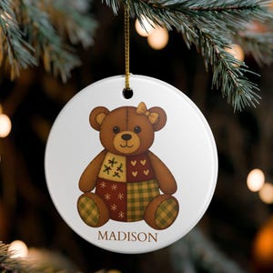 Pode incluir: Enfeite de Natal redondo e branco com um desenho de urso de pelúcia marrom. O urso tem um padrão de patchwork em tons de marrom, verde e vermelho. O nome "MADISON" é impresso na parte inferior. Uma corda dourada é anexada para pendurar.