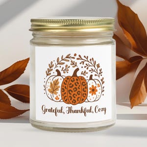 Könnte beinhalten: Ein Glas-Kerzenglas mit goldenem Deckel. Das Etikett zeigt drei Kürbisse mit herbstlichen Designs und dem Text "Grateful, Thankful, Cozy". Die Kürbisse sind von einem dekorativen floralen Design umgeben. Die Kerze ist ein Produkt zum Thema Herbst.