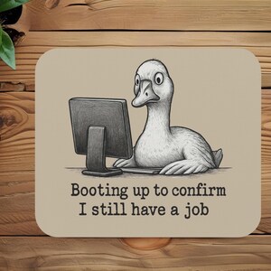 May include: A beige mousepad features a cartoon duck looking at a computer screen. The text reads, "Booting up to confirm I still have a job." The duck is white with a black and white computer screen.
