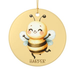 May include: A round, yellow ceramic ornament featuring a cartoon bee with a smiling face, black and yellow stripes, and light blue wings. The name "HARPER" is printed in brown letters at the bottom. A gold string is attached for hanging.