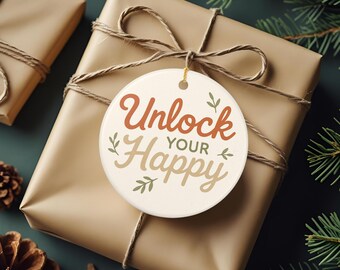 May include: A round white ceramic ornament featuring the text "Unlock YOUR Happy" in brown and tan script lettering, accented with small green leaf illustrations. The ornament is tied with a thin gold string to a brown gift box wrapped in twine.