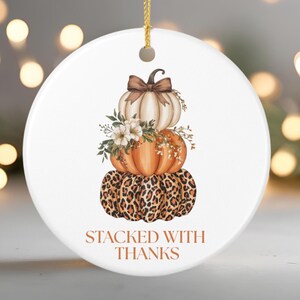 Könnte beinhalten: Runder weißer Keramikschmuck mit einem gestapelten Kürbismotiv. Der obere Kürbis ist weiß, der mittlere ist orange und der untere hat einen Leopardenmuster. Er hat eine goldene Schnur und den Text "STACKED WITH THANKS".