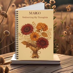 May include: A spiral-bound notebook with the name "MARGO" and the phrase "Gathering My Thoughts" printed on the cover. The cover features a bouquet of autumn-colored flowers, including sunflowers and dahlias. The notebook rests on a stack of books.