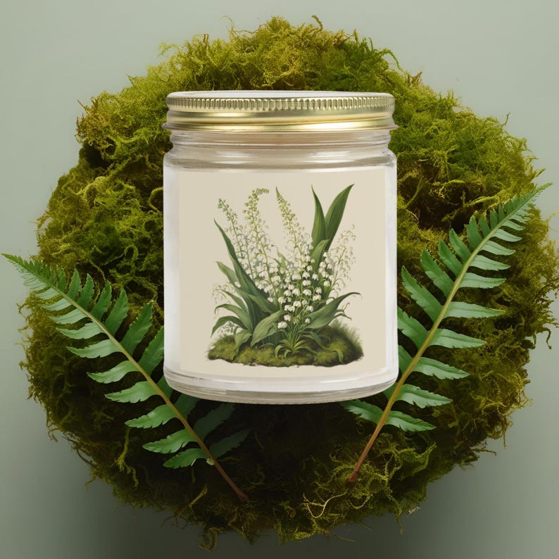 May include: A glass jar candle with a gold lid, featuring a botanical illustration of lily of the valley flowers. The candle sits on a bed of green moss, with fern leaves on either side. The candle label has a beige background.