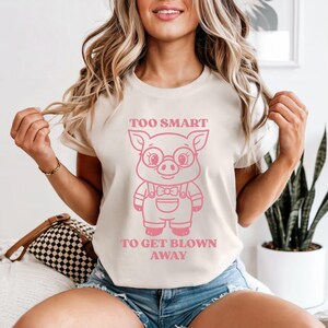 May include: A cream-colored t-shirt featuring a pink cartoon pig wearing glasses and overalls. The text "TOO SMART TO GET BLOWN AWAY" is printed above and below the pig illustration. The shirt has short sleeves.