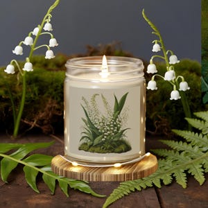 May include: A lit candle in a clear glass jar featuring a lily of the valley botanical illustration. The candle rests on a wooden coaster with fairy lights, surrounded by greenery and artificial lily of the valley.