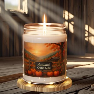 Könnte beinhalten: Eine brennende Kerze in einem Glas mit einem Etikett, das eine Herbstlandschaft mit Kürbissen und dem Text "Autumn's Quiet Side" zeigt. Die Kerze steht auf einem Holzuntersetzer.