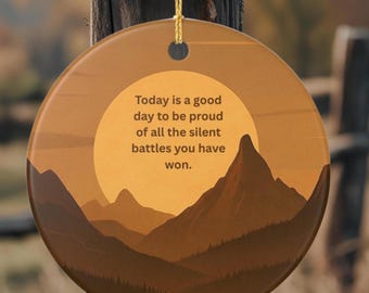 May include: A round ceramic ornament featuring a sunset mountain landscape in shades of brown and orange. The text reads: Today is a good day to be proud of all the silent battles you have won. A gold string is attached to the top for hanging.