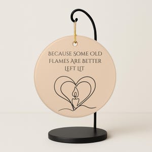 May include: A round, beige ceramic ornament with the text "BECAUSE SOME OLD FLAMES ARE BETTER LEFT LIT" and a black line drawing of a candle inside a heart. The ornament hangs from a gold cord on a black metal stand.