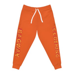Puede incluir: Pantalones deportivos naranjas con cordón blanco. El texto "BAGNABY" está impreso verticalmente en la pierna izquierda y "CLOTHING" en la pierna derecha, ambos en una fuente con forma de llama.
