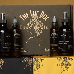 Può includere: Un'esposizione di prodotti per la cura dei capelli di "The Loc Doc Essentials". Il set include flaconi ambrati scuri di olio per la crescita dei capelli, spray idratante e una scatola con il logo del marchio. Le bottiglie sono etichettate con nomi e dimensioni dei prodotti.