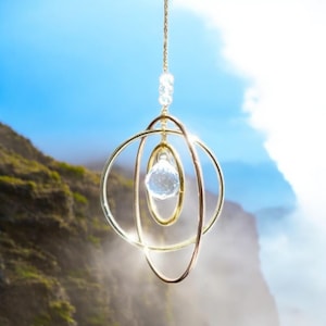 May include: A hanging gold-colored wind chime with a clear crystal ball and three concentric rings. The wind chime is suspended from a delicate chain with small, clear beads. The background features a blurred view of a mountain and sky.