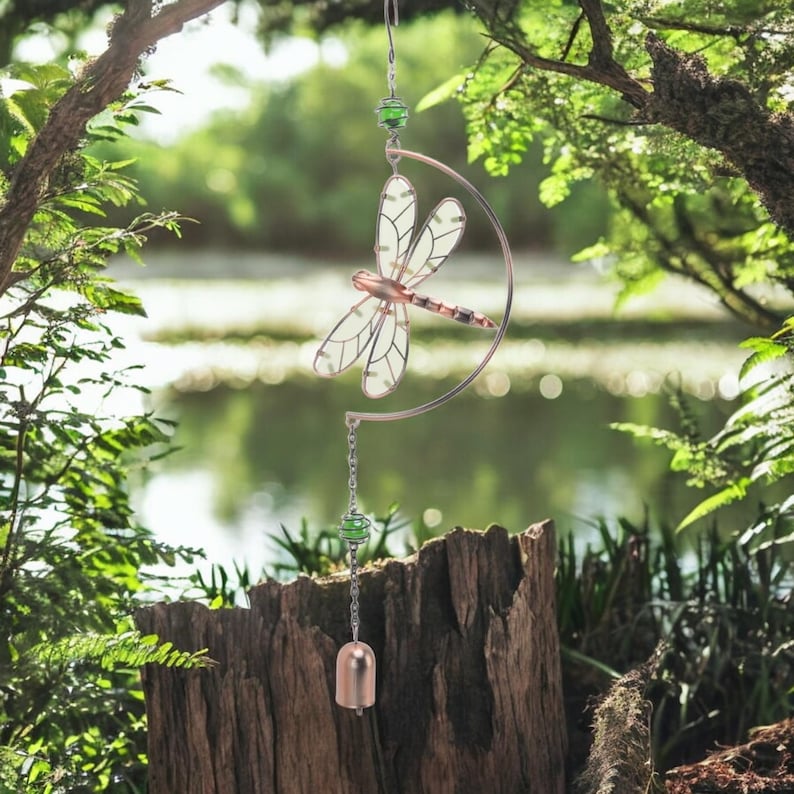 May include: A decorative dragonfly wind chime. The dragonfly has a copper-colored body and wings with a clear, patterned design. The chime hangs from a curved metal frame with green beads and a small bell.
