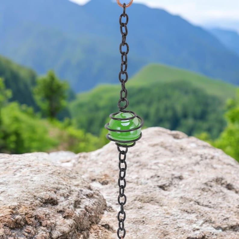 May include: A decorative rain chain with a green glass ball encased in a black metal spiral. The chain is made of black metal links and is suspended from a copper ring. The background features a blurred view of mountains and greenery.