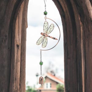 May include: A decorative dragonfly wind chime. The dragonfly has translucent wings and a copper-colored body, suspended from a copper-colored crescent moon. Green beads and a small bell hang below. The chime is viewed through a wooden archway.