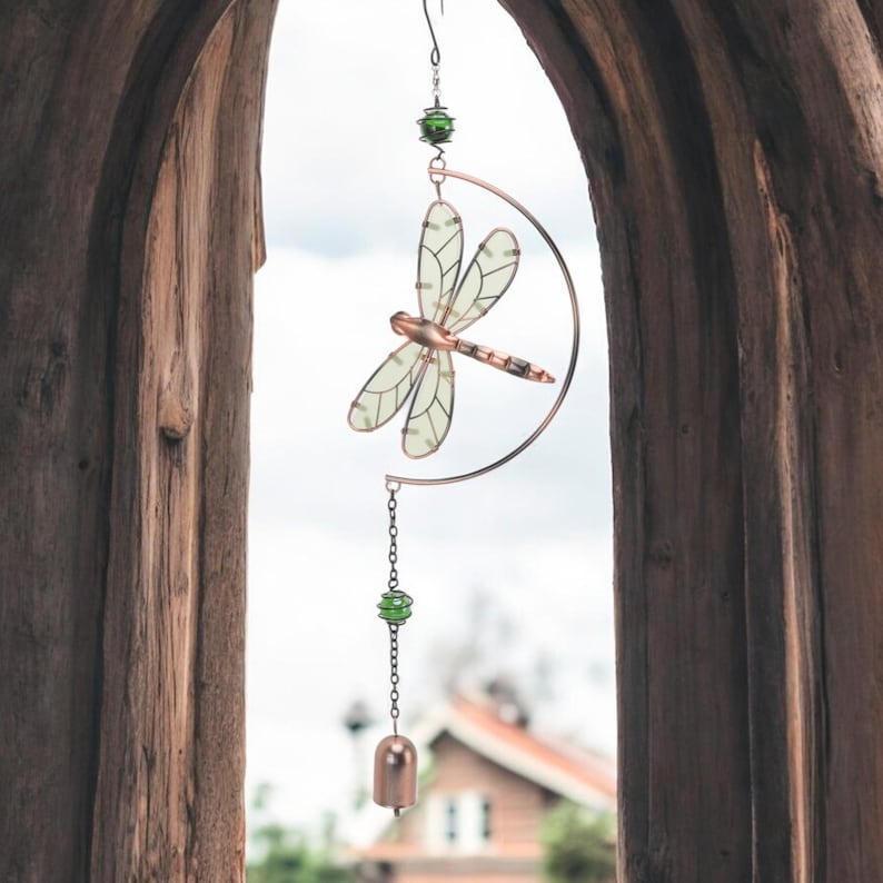 May include: A decorative dragonfly wind chime. The dragonfly has translucent wings and a copper-colored body, suspended from a copper-colored crescent moon. Green beads and a small bell hang below. The chime is viewed through a wooden archway.