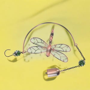May include: A copper-colored dragonfly wind chime with clear wings and black outlines. The dragonfly is suspended from a curved wire, with green glass beads and a small bell at the bottom. The background is a solid yellow.