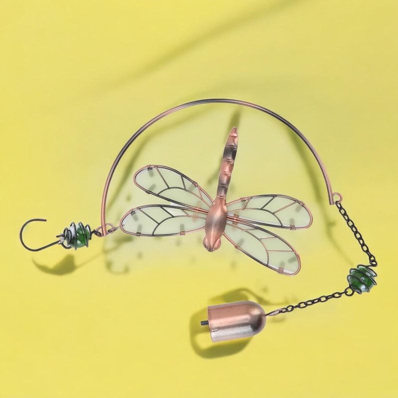 May include: A copper-colored dragonfly wind chime with clear wings and black outlines. The dragonfly is suspended from a curved wire, with green glass beads and a small bell at the bottom. The background is a solid yellow.