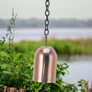 May include: A copper-colored bell hanging from a black chain. The bell has a rounded top and a flared bottom. The background features green foliage and a body of water, creating a natural setting.